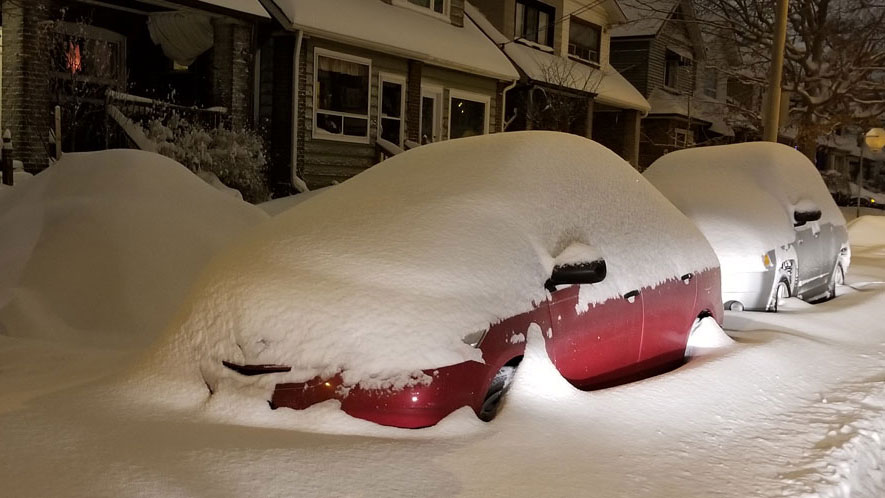 Cars covered in a thick blanket of snow are parked on a snow-covered residential street.